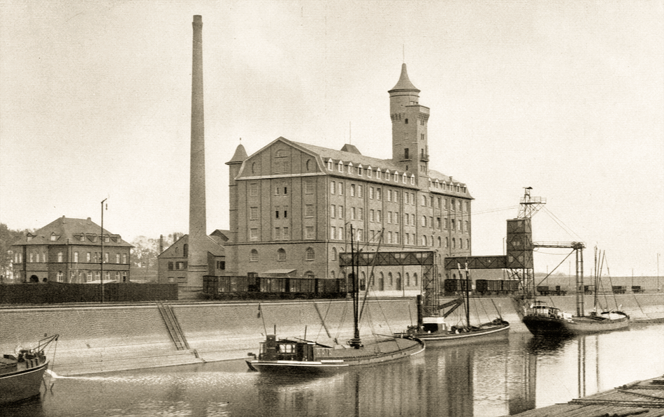Historische Aufnahme des Deutzer Hafens: mehrgeschossiger Mühlenbau mit Eckturm und hohem Schornstein an der Kaimauer, davor Lastkähne im Hafenbecken und Güterwagen auf dem Kai